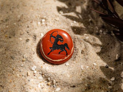 Runder Handschmeichler mit schwarzem Schütze-Motiv liegt auf Sand