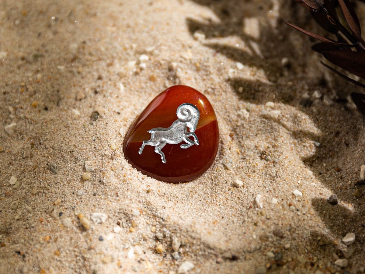 Stimmungsbild-gerundeter-Sternzeichen-Handschmeichler-Motiv-Widder Rotbrauner Taschenstein mit Widder-Motiv liegt auf Sand